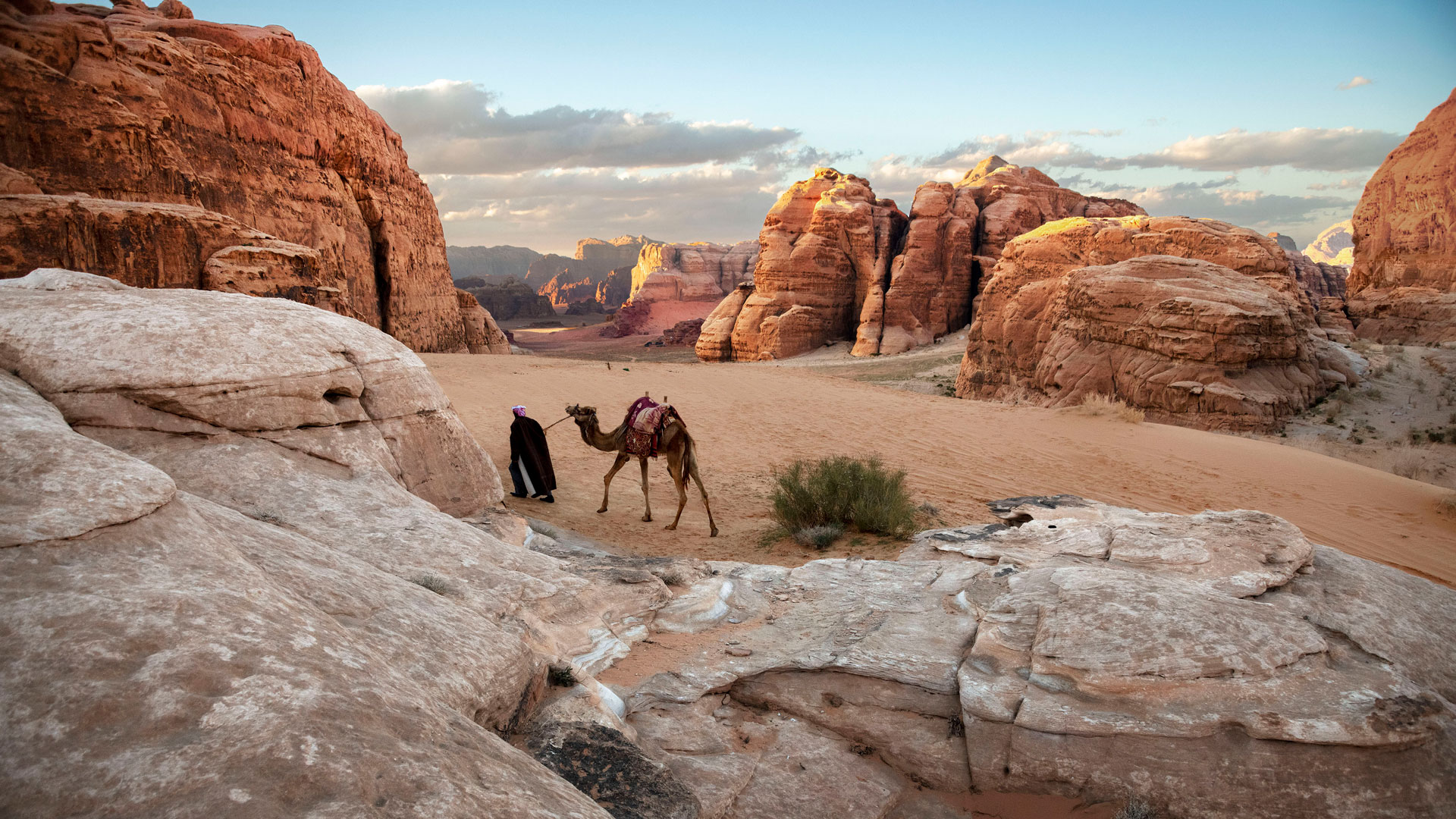 Une scène de désert où un homme mène un chameau à travers un paysage rouge et rocheux.