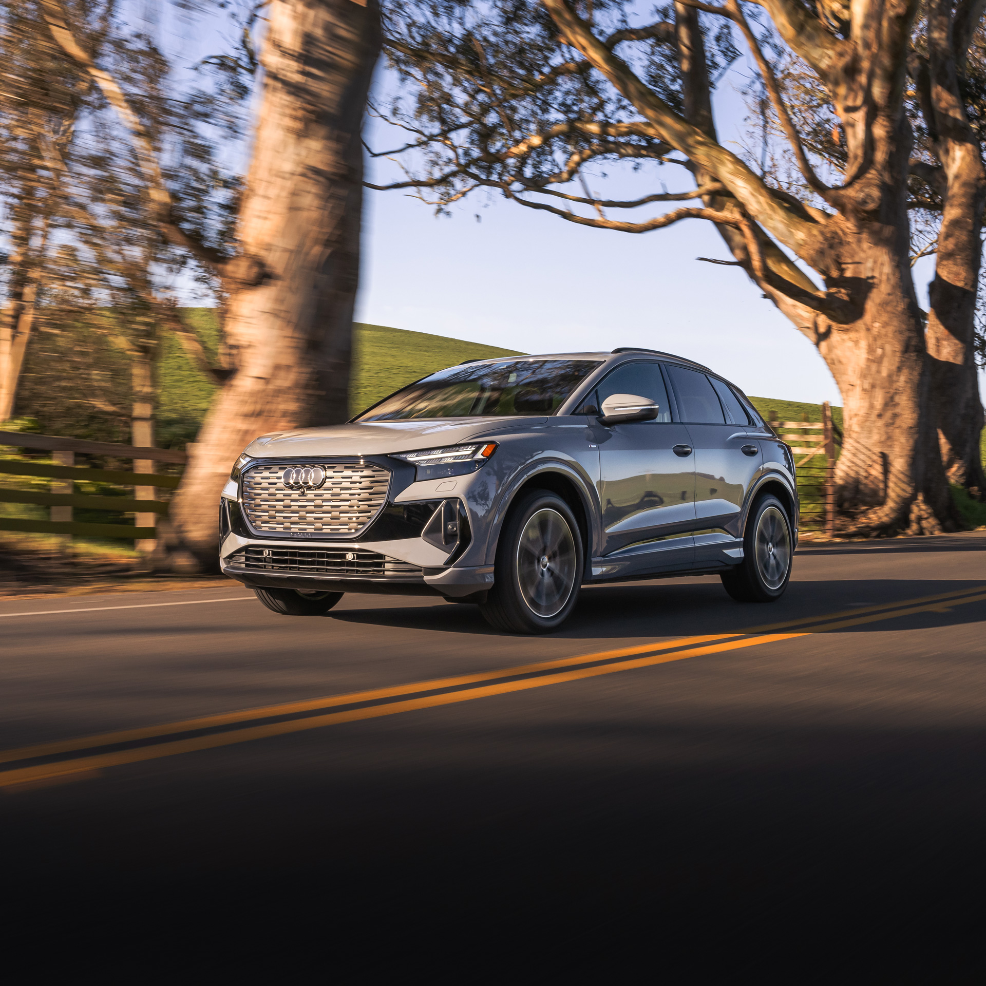 Three-quarter front view of a grey Audi Q4 Sportback e-tron driving on a tree-lined row. 